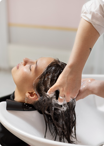 La imagen muestra a una persona recibiendo un lavado de cabello en un salón de belleza. El estilista está aplicando shampoo y masajeando el cuero cabelludo de la cliente mientras ella está reclinada, disfrutando del servicio. El ambiente parece relajante y cuidado, lo que hace de este momento un proceso de relajación y cuidado personal.