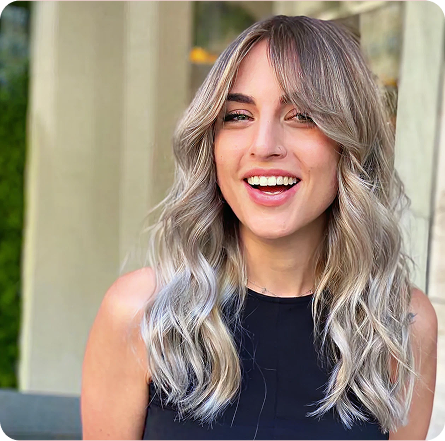 La imagen muestra a una mujer con un estilo de cabello largo y ondulado, con un tono rubio platino y un suave degradado. Su corte lleva flequillo que enmarca su rostro de manera natural, aportando frescura y luminosidad. La melena tiene mucho volumen y movimiento, lo que le da un aire relajado pero sofisticado. Este look es ideal para quienes buscan un estilo moderno y versátil, que resalta con facilidad.