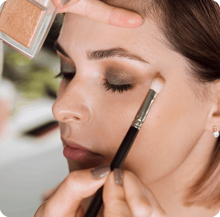 La imagen muestra a una persona recibiendo un maquillaje profesional. El enfoque está en la aplicación de sombra de ojos con una brocha, mientras el maquillador trabaja cuidadosamente en los detalles para realzar la mirada. El maquillaje resalta los ojos con tonos metálicos, mientras que el resto de su rostro tiene un acabado natural y fresco. La atención al detalle refleja una técnica experta y precisa, ideal para un look elegante y sofisticado.