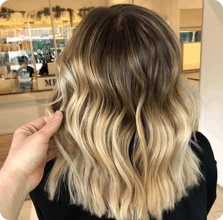 La imagen muestra un corte de cabello bob con ondas suaves, con un degradado de color que va desde un tono oscuro en las raíces hasta un rubio cálido en las puntas. Las ondas añaden movimiento y textura al cabello, dando un look natural y vibrante. Este estilo es perfecto para quienes buscan un corte fresco, moderno y de fácil mantenimiento, que resalta la luminosidad del cabello.