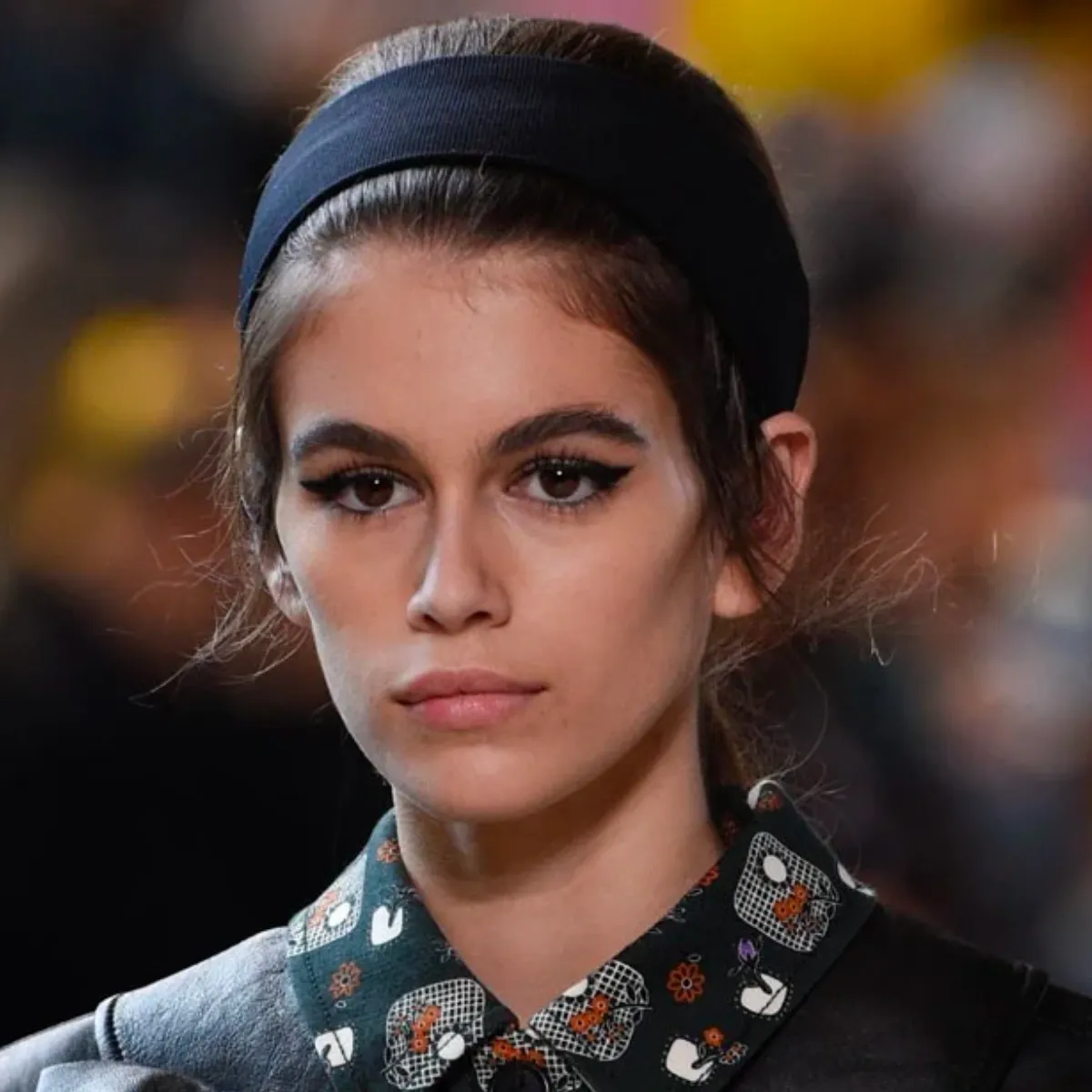 La imagen muestra a una mujer con un look elegante y sofisticado. Lleva una diadema negra que complementa perfectamente su peinado recogido. El maquillaje resalta con un delineado de ojos marcado, mientras que su camisa de cuello alto con detalles florales le da un toque distintivo. El conjunto tiene un aire vintage y moderno a la vez, ideal para una ocasión especial.