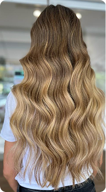 La imagen muestra una melena larga y ondulada con un tono rubio cálido que combina varias tonalidades para un efecto degradado. Las ondas suaves aportan volumen y movimiento al cabello, creando un look natural y voluminoso. Este estilo transmite frescura y vitalidad, ideal para quienes buscan un toque relajado y sofisticado.