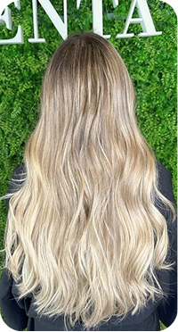 La imagen muestra un cabello largo con ondas suaves y un tono rubio degradado que va aclarando hacia las puntas. El cabello tiene un acabado natural y fluido, con mucho movimiento, lo que le da un toque fresco y relajado. El fondo verde resalta la melena, creando un contraste que enfatiza la luminosidad y la textura del cabello. Es un look ideal para quienes buscan un estilo elegante pero con un aire desenfadado.