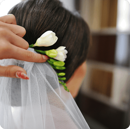 La imagen muestra a una persona colocando flores blancas en el peinado de una mujer, probablemente como parte de un peinado de novia. Se observa que la floristería se coloca cuidadosamente, mientras que la mujer parece estar lista para una ocasión especial. La escena refleja la atención al detalle y el cuidado que se pone en los peinados para eventos importantes.