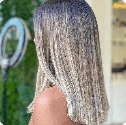 La imagen muestra un peinado lacio y elegante con un color de cabello rubio degradado, que va de un tono oscuro en la raíz a un rubio claro en las puntas. El cabello está perfectamente peinado, reflejando un acabado suave y brillante. La imagen destaca la transición del color y la salud del cabello.