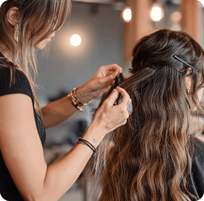La imagen muestra a una estilista trabajando en el cabello largo y ondulado de una clienta. Está usando un peine o pinza para sujetar una sección del cabello mientras probablemente lo prepara para un peinado o tratamiento. El ambiente del salón tiene una luz cálida y un fondo desenfocado, lo que da una sensación de acogimiento y confort.