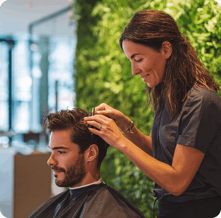 La imagen muestra a un estilista trabajando en el cabello de un hombre en un salón de belleza. La estilista está cuidadosamente peinando o cortando el cabello del cliente, quien parece relajado y cómodo. El fondo de la imagen está decorado con una pared verde, probablemente de plantas, lo que aporta una atmósfera natural y relajante al entorno del salón.