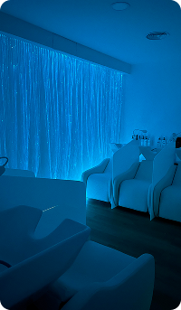La imagen muestra un salón con una iluminación ambiental azul que resalta una serie de sillas blancas. Al fondo, hay cortinas que también tienen un tono suave, creando una atmósfera relajante y moderna. El diseño minimalista sugiere un espacio de estética o bienestar.