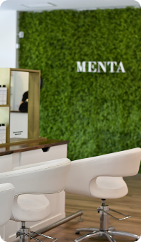 La imagen muestra un moderno salón de belleza con dos sillas blancas y una pared de plantas verdes. En la pared se encuentra el nombre "MENTA" en letras grandes, lo que sugiere que es un espacio dedicado al cuidado personal. El ambiente tiene un estilo minimalista y elegante.