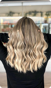 La imagen muestra a una persona con cabello largo y ondulado, con un degradado de colores que va desde un tono oscuro en las raíces hasta un rubio claro en las puntas. La persona está mirando hacia atrás, con las manos sujetando su cabello, en un entorno luminoso.