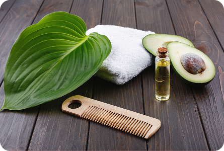 La imagen muestra una escena de cuidado personal, con una toalla blanca, un peine de madera, una hoja verde grande, medio aguacate y un frasco de aceite, sobre una superficie de madera oscura. Los elementos evocan una sensación de naturalidad y bienestar.