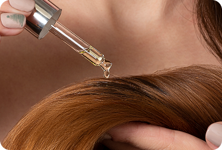 La imagen muestra una mano aplicando un aceite o suero capilar sobre el cabello. El foco está en la gota de aceite que cae sobre el cabello, con un fondo desenfocado, resaltando la textura del cabello.