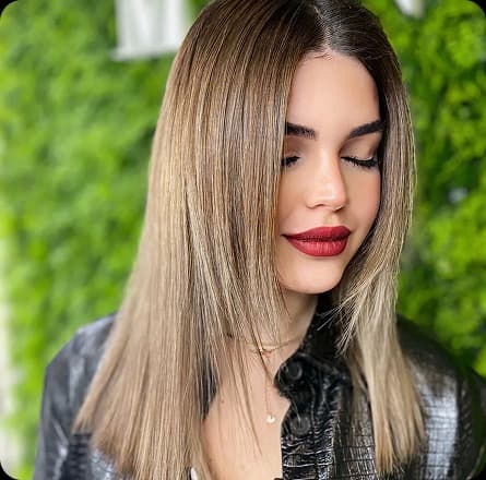 La imagen muestra a una mujer con un corte de cabello lacio y elegante, con un tono de rubio suave y un acabado brillante. Su look está complementado con un maquillaje natural y un estilo moderno. El fondo verde resalta el contraste del cabello, creando una atmósfera fresca y sofisticada.
