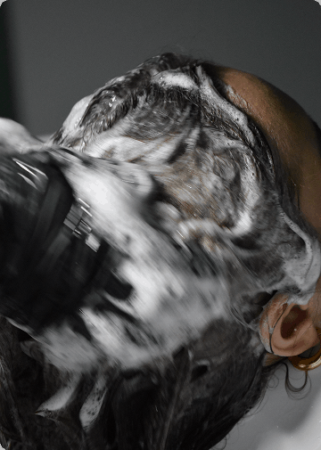 La imagen muestra el proceso de aplicación de champú en el cabello de una persona, con espuma visible cubriendo el cuero cabelludo y el cabello. El enfoque en el champú y la espuma sugiere que se está realizando un tratamiento o lavado detallado. El fondo está difuso, lo que destaca el cabello y el champú en primer plano, creando una sensación de enfoque en el cuidado capilar.