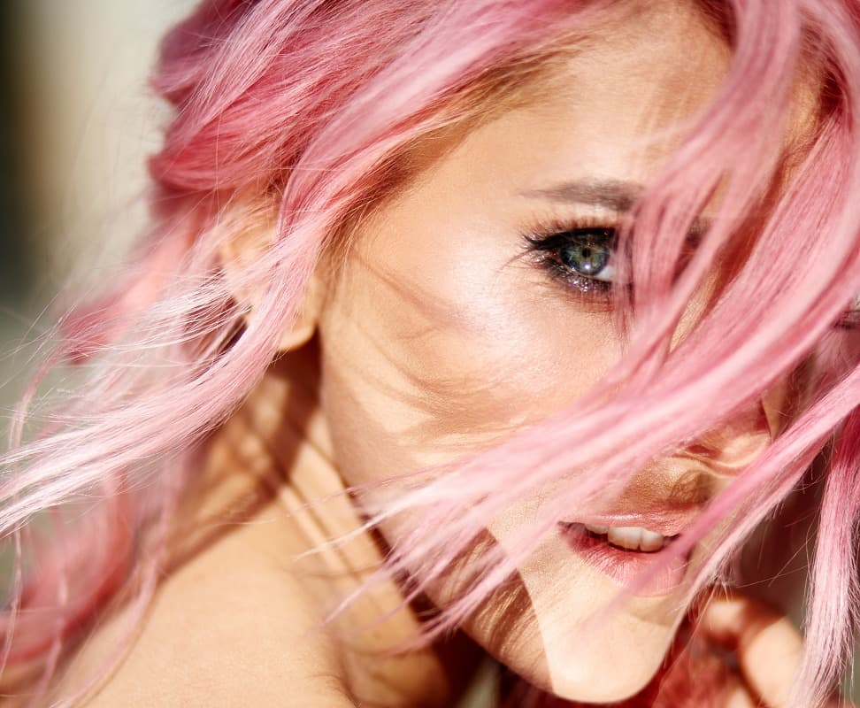 La imagen muestra a una mujer con cabello rosa, ligeramente al viento, que cubre parcialmente su rostro. Ella tiene una mirada intensa, con los ojos enfocados y una leve sonrisa, mientras la luz resalta sus rasgos y cabello