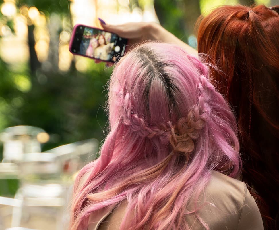 La imagen muestra a dos personas, una con el cabello rosa y trenzado, y la otra con el cabello rojizo, mientras se toman una selfie con un teléfono móvil. El fondo muestra un entorno natural con árboles y sillas.